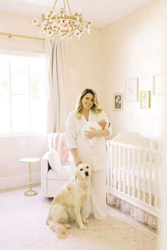 A woman holding a baby stands beside a crib in a softly lit nursery, with a large dog sitting at her feet.