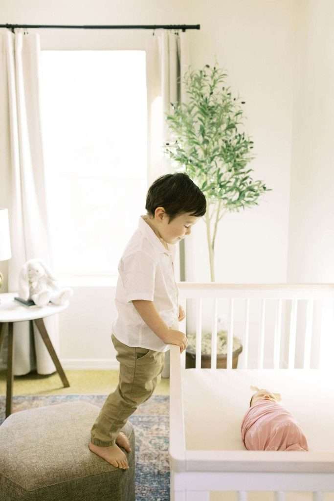 A young boy stands on a stool next to a crib, looking at a swaddled baby lying inside. A potted plant and a curtained window are visible in the background.
