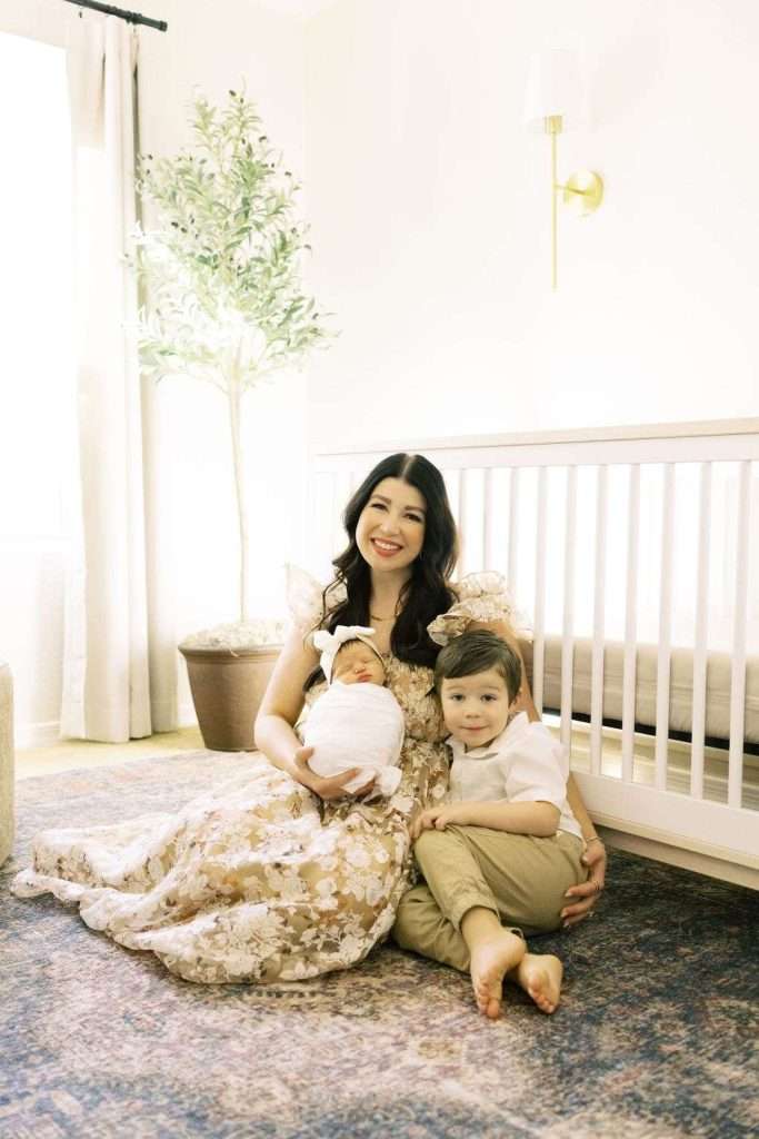 Woman sitting on a rug in a nursery, holding a swaddled baby and sitting next to a young child in front of a crib and potted plant.
