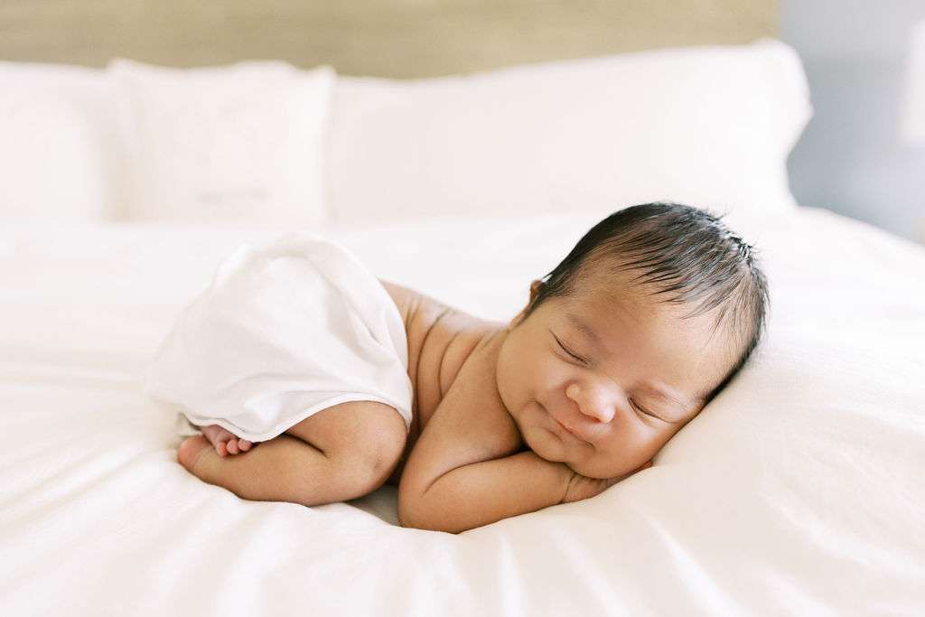 Newborn baby sleeping on a white bed, wrapped loosely in a white blanket, with head resting on arms and eyes closed.