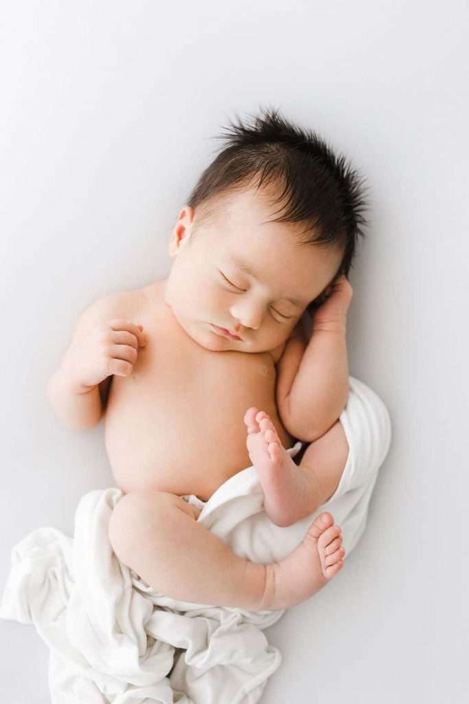 A newborn baby with dark hair sleeps on a white blanket, one hand near the head and legs slightly bent, with a peaceful expression.