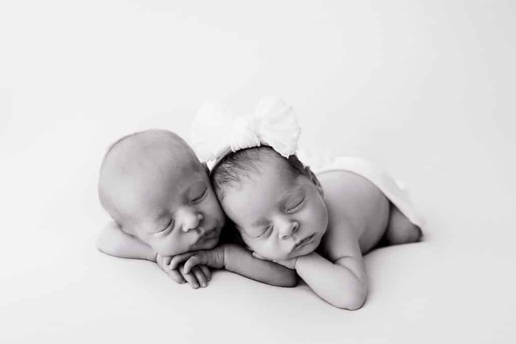 Two newborn babies lie side by side on a soft surface, both sleeping peacefully; one baby wears a large white bow on their head.