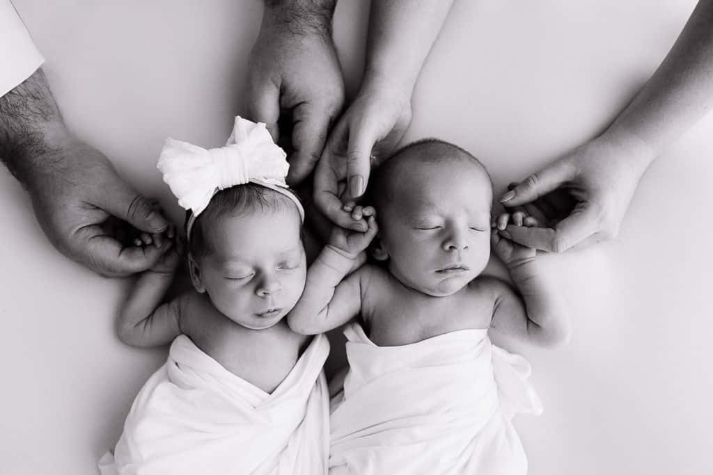 Two newborn babies swaddled in blankets lay side by side, each holding the hands of adults. One baby wears a headband with a bow. The image is black and white.