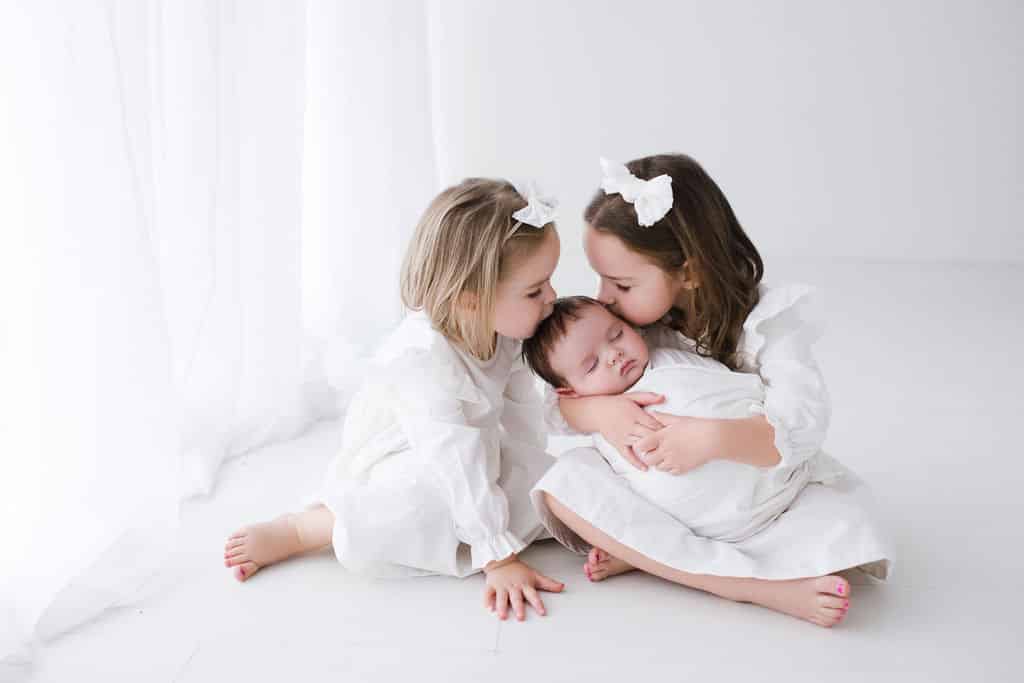 Two young girls in white dresses sit on the floor, gently embracing and kissing a sleeping baby swaddled in white, against a bright white background.