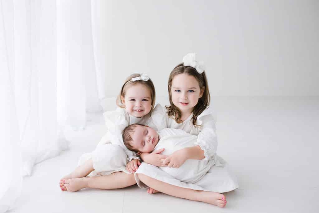 Two young girls in white dresses sit on the floor, smiling, while holding a swaddled baby in their laps. All three children wear white bows in their hair.