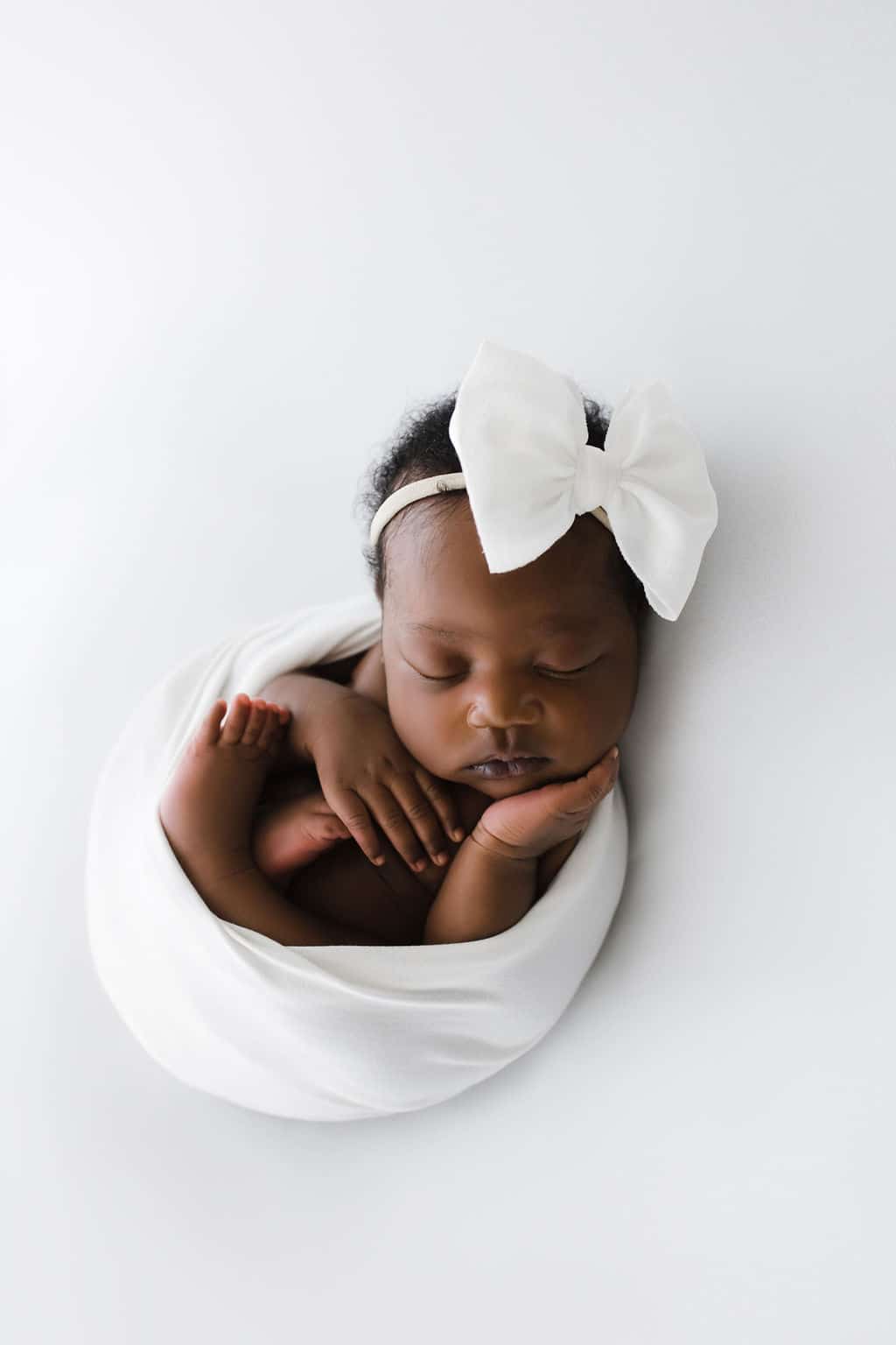 Newborn baby swaddled in a white blanket, sleeping with hands under chin, wearing a white headband with a large bow, on a white background.