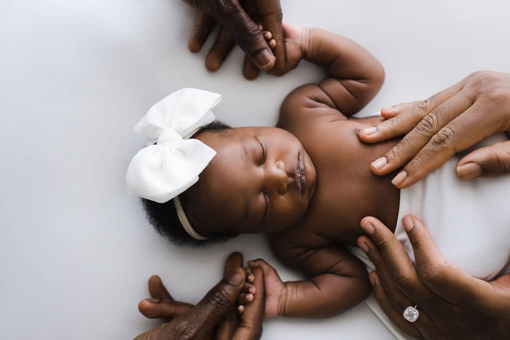 Newborn baby with a white bow headband lies on a white surface, surrounded by gentle adult hands touching her arms and holding her hands.