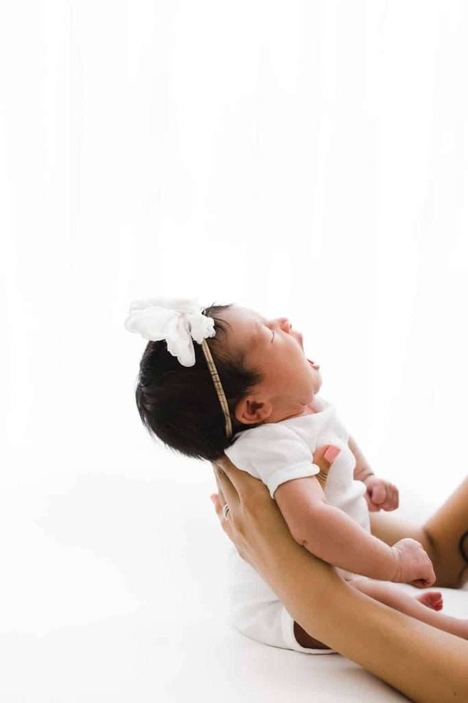 A newborn baby wearing a white headband with a bow is being held upright by an adult on a white background.