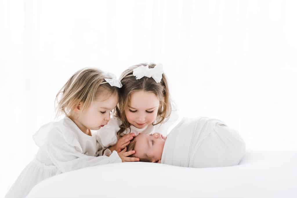 Two young girls in white outfits gently touch and look at a newborn baby swaddled in white on a white background.