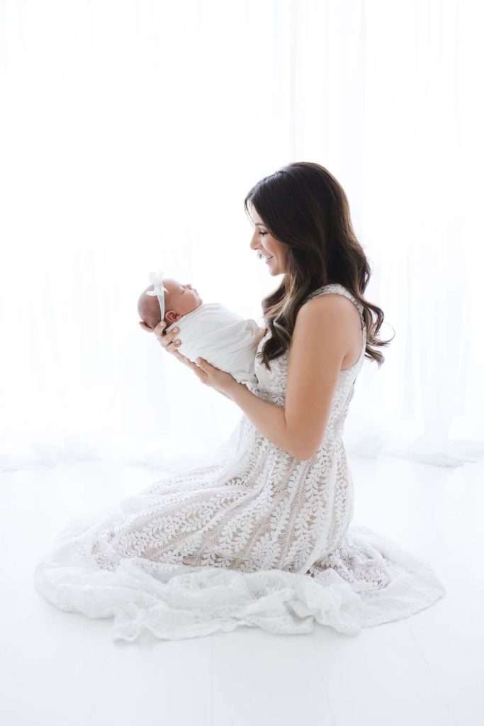 A woman in a white lace dress kneels on the floor, smiling and holding a swaddled newborn baby in front of a bright, white background.