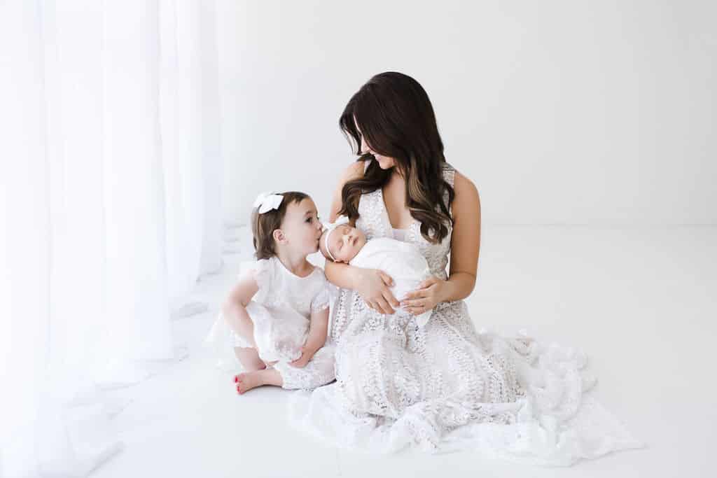 A woman in a white dress holds a swaddled newborn while a young girl in white kisses the baby’s head. They are seated on a white floor with sheer curtains in the background.