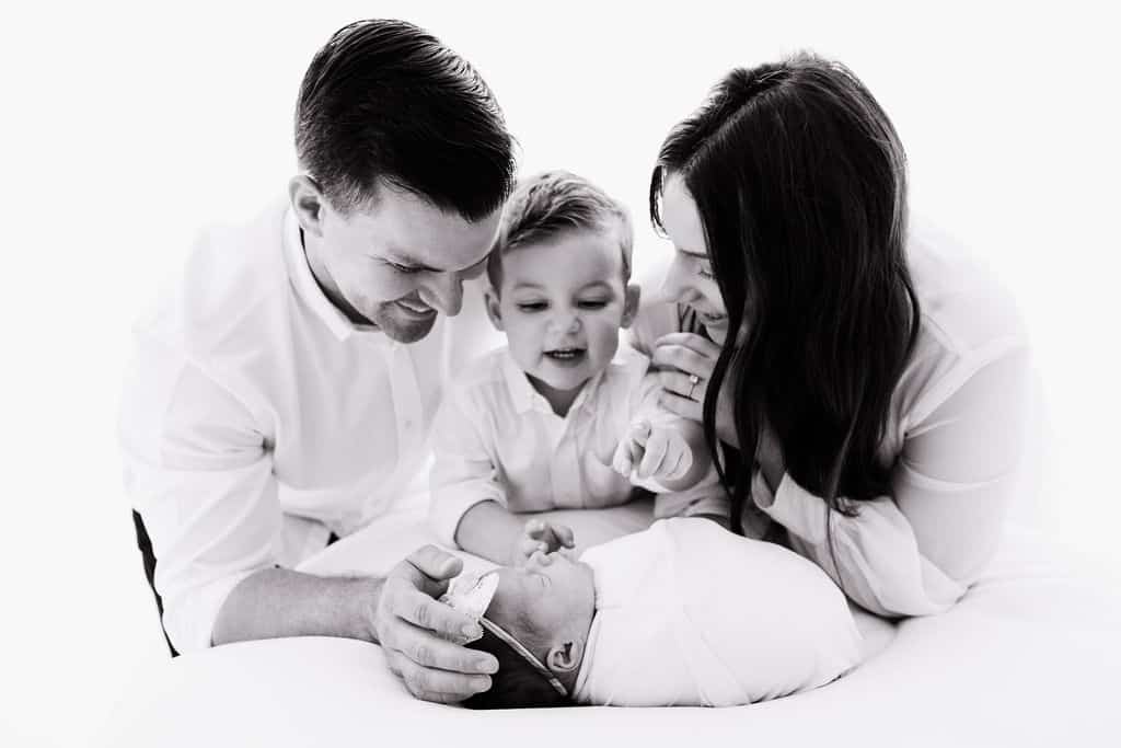 A family of four, dressed in white, smiles and looks at a swaddled newborn while gathered closely together on a white surface.