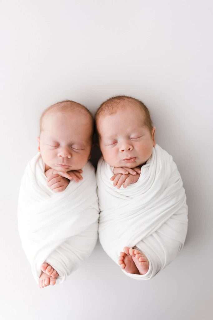 Two newborn babies wrapped in white blankets lie side by side on a white background, both sleeping peacefully with their heads touching.