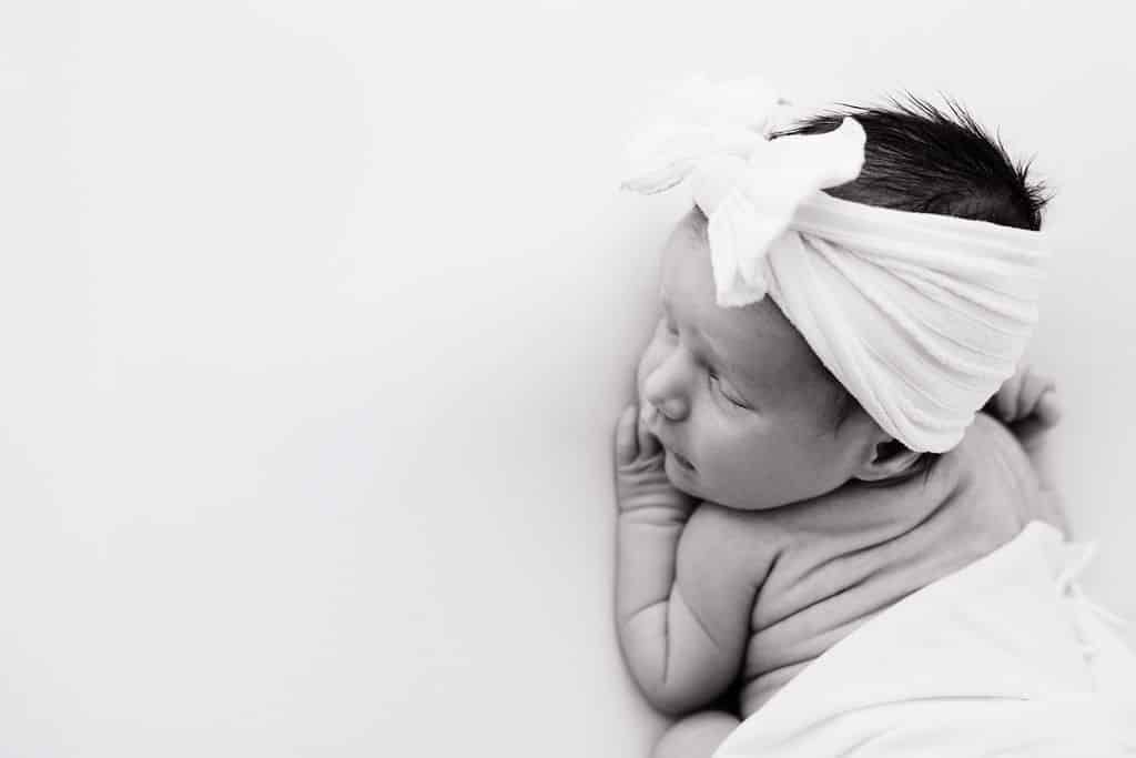 Black and white photo of a baby lying on their side, wrapped in a blanket, wearing a headband with a large bow, looking slightly upward.