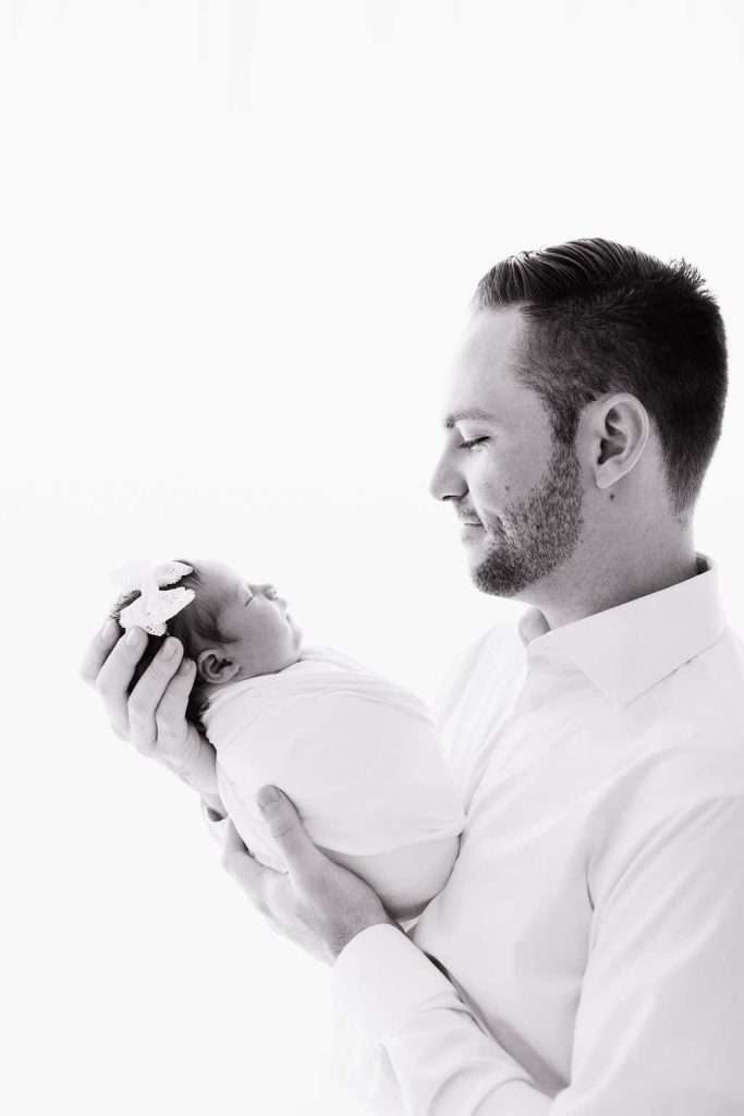 A man in a light shirt gently holds a swaddled newborn with a bow headband, both facing each other against a white background.