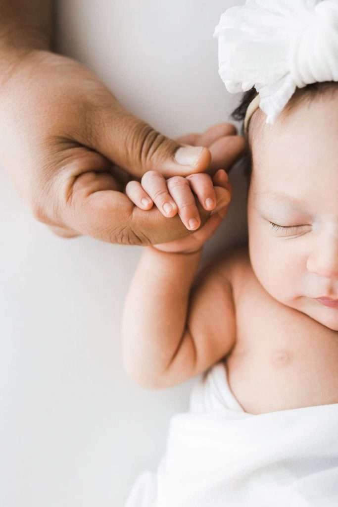 Close-up of an adult hand gently holding a newborn baby’s hand; the baby wears a white headband and is wrapped in a white blanket.