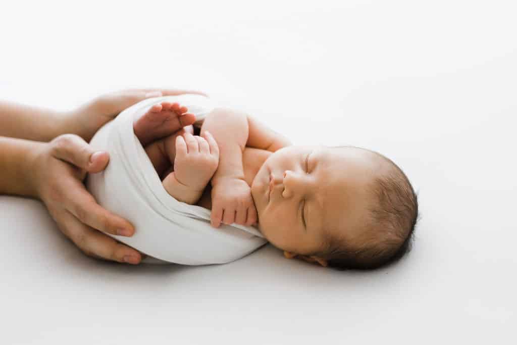 A newborn baby wrapped in a white blanket sleeps on a white surface while being gently held by an adult's hands.