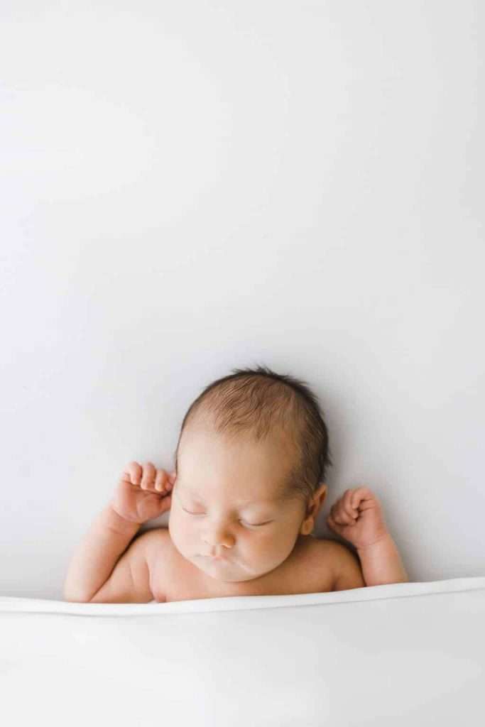 A newborn baby lies on a white surface, eyes closed and arms raised, partially covered by a white blanket.