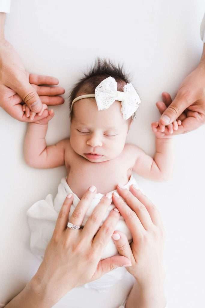 Newborn baby with a white bow headband lies on a white surface, holding hands with two adults and gently touched by another adult's hands.