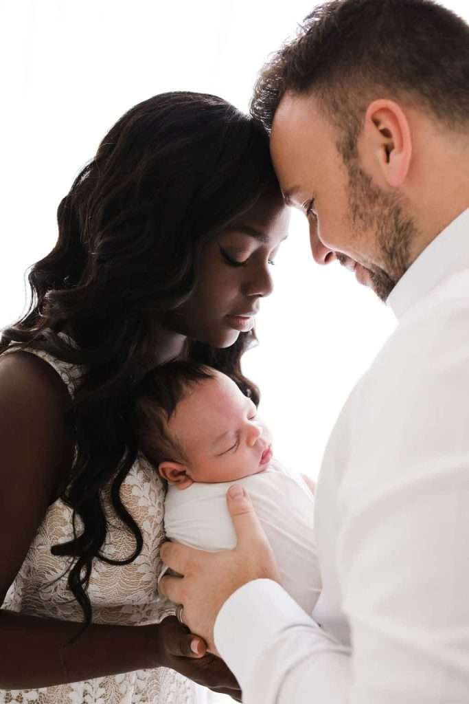 A woman and man gently hold a sleeping newborn wrapped in a white blanket, standing close with their foreheads touching.
