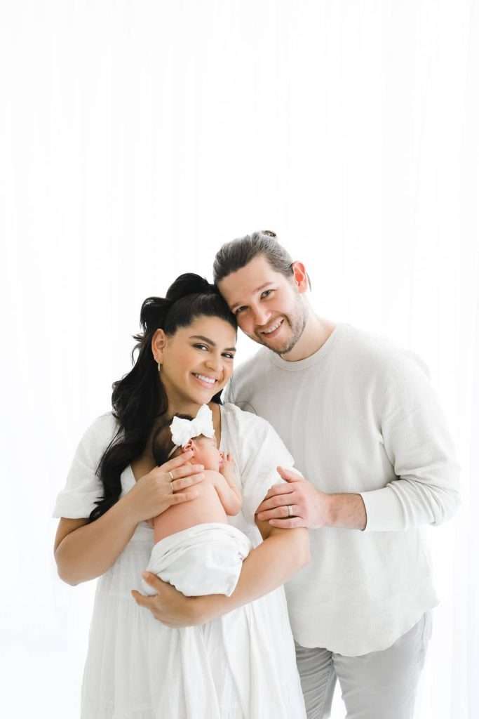 A woman holds a newborn wrapped in a white blanket. A man stands beside her, both adults smiling at the camera against a white background.