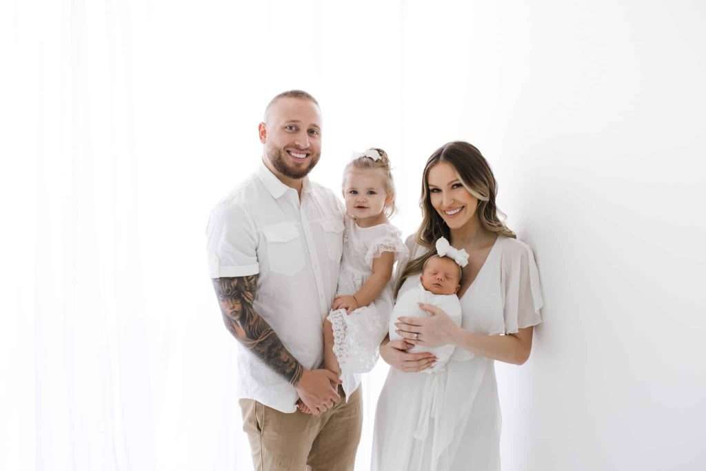 A man and woman stand together, smiling, with a young girl in the man's arms and a baby wrapped in a blanket in the woman's arms, all dressed in light clothing against a white background.