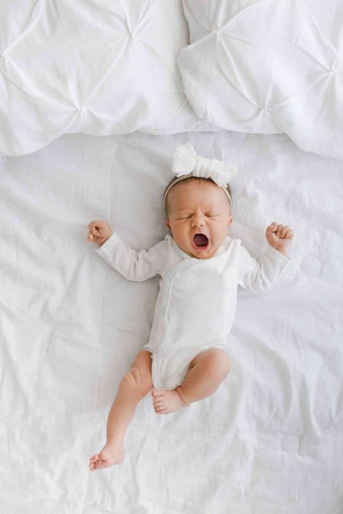 Baby in a white onesie with a white bow headband lying on a white bed, yawning with arms raised.