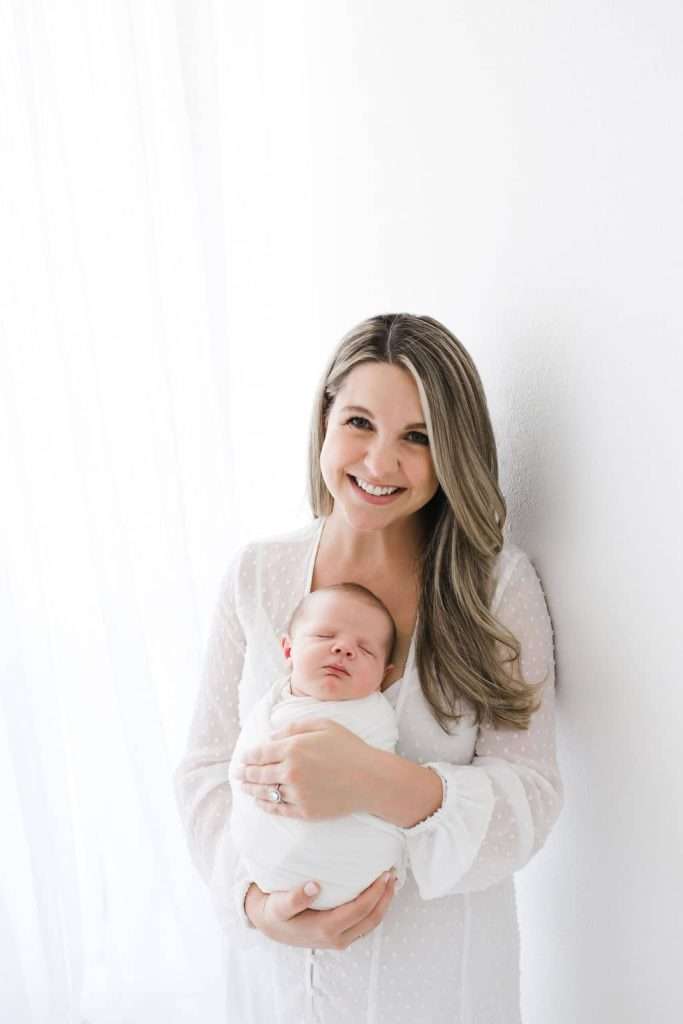 Woman in a white dress stands by a white wall, smiling and holding a swaddled newborn baby in her arms.