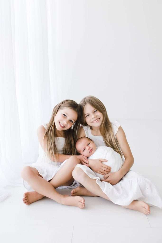 Two young girls in white dresses sit on the floor, smiling and holding a baby wrapped in a white blanket, against a bright white background.