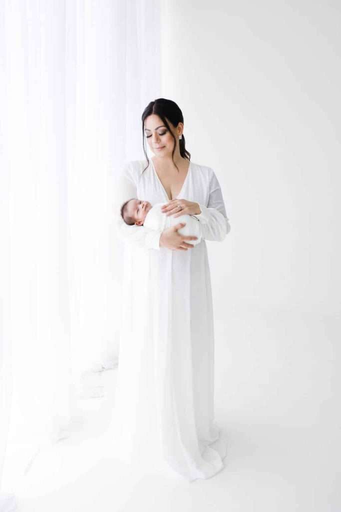 Woman in a long white dress stands in a bright, white room, gently holding a swaddled baby in her arms and looking down at the infant.