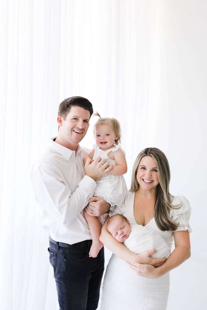 A family of four poses together. The father holds a toddler girl, while the mother holds a newborn. All wear white and are smiling against a white background.