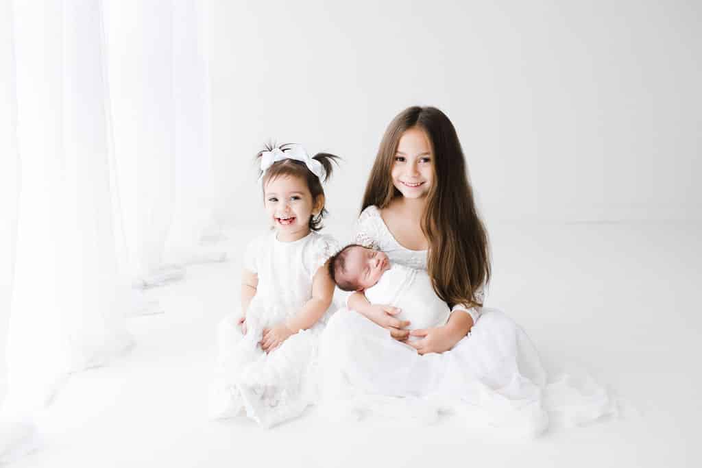 Three young children in white clothes sit on a white floor; a girl with long hair holds a baby, and a toddler sits beside them, smiling at the camera.