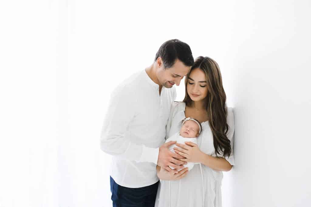 A man and a woman dressed in white gently hold a swaddled newborn, standing close together against a plain white background.