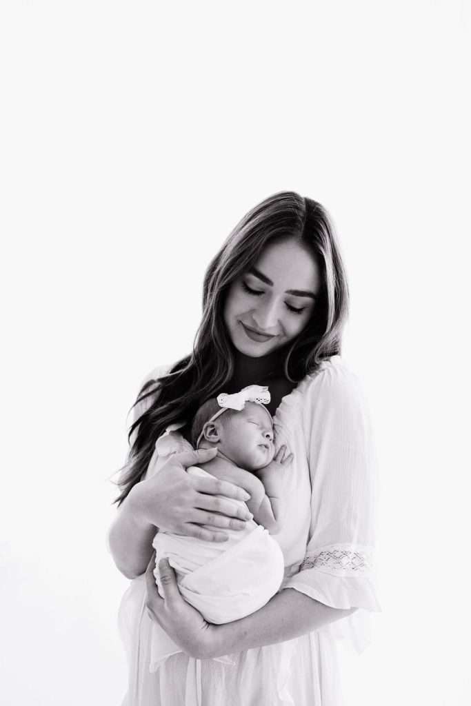 Woman in a white dress holds a sleeping newborn wrapped in a blanket, both facing forward, against a white background.