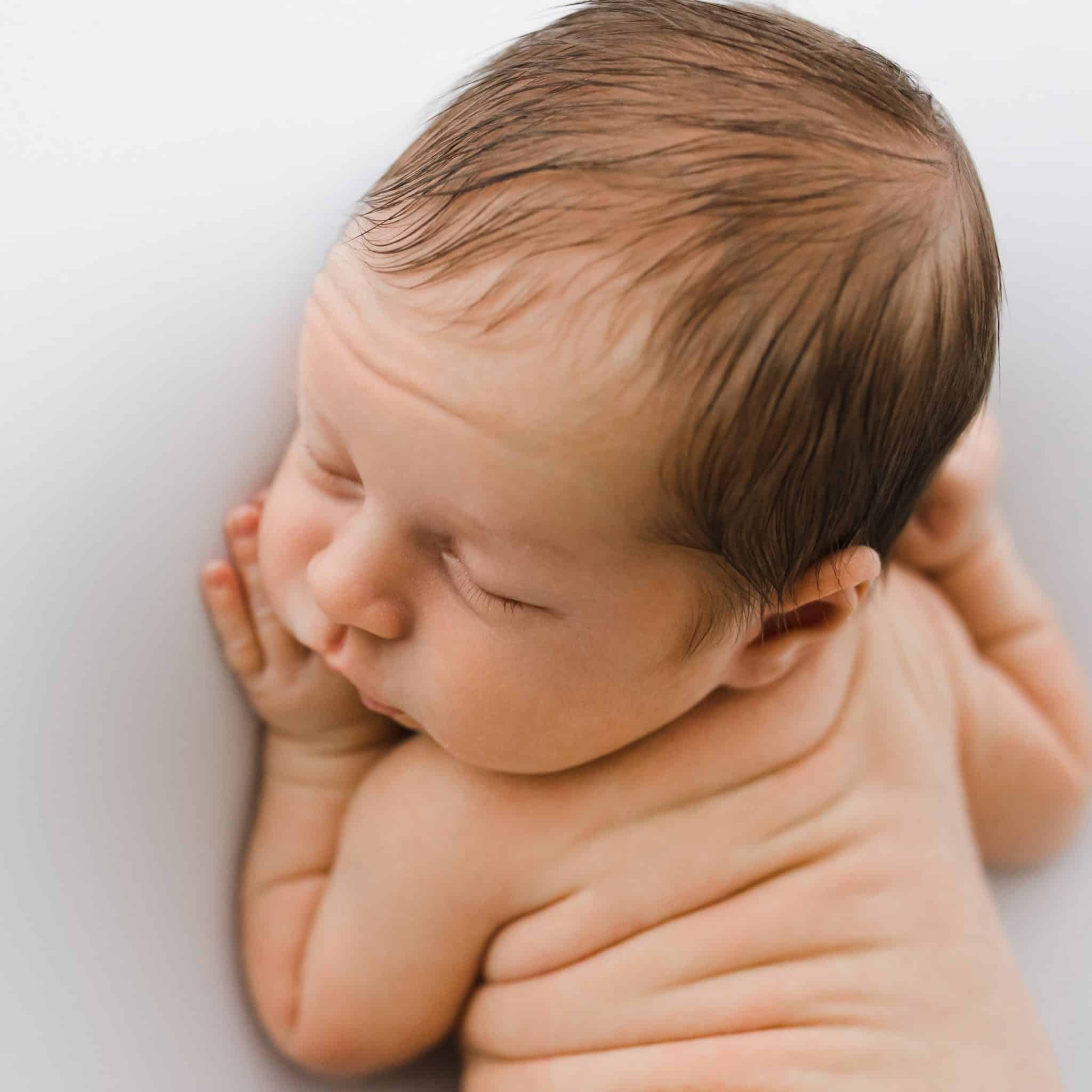 A newborn baby lies on their stomach with eyes closed, resting one hand against their cheek on a white background.