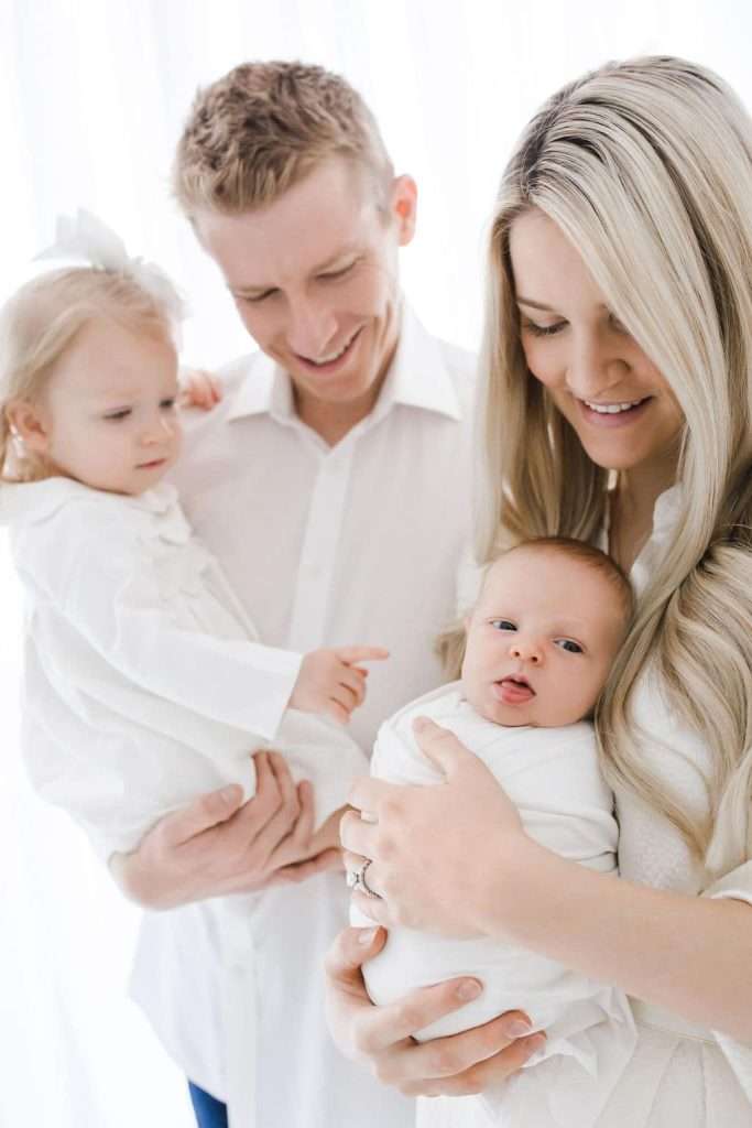 A man holds a young girl while a woman holds a swaddled baby. All four wear white clothing and stand closely together, smiling.