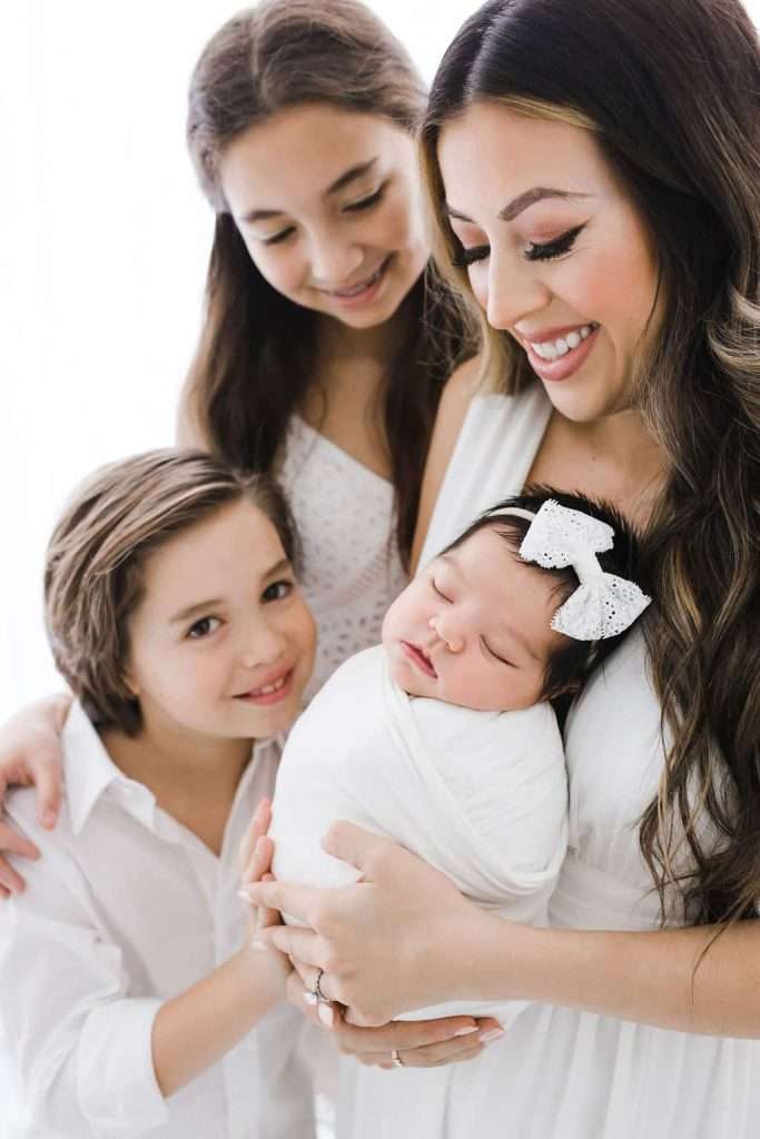Woman holding a swaddled baby with a bow headband, standing next to a smiling boy and girl, all wearing white.