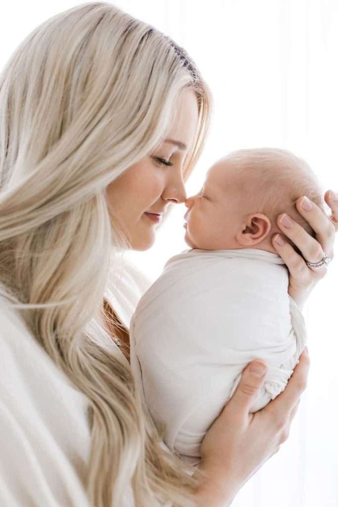 Woman holding a swaddled newborn, touching noses and smiling in a softly lit, white background setting.