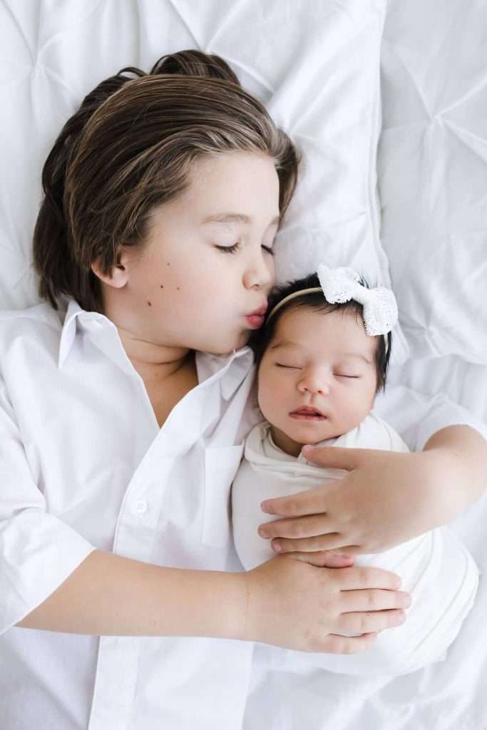 A young boy in a white shirt lies on a bed, gently kissing the forehead of a swaddled baby wearing a white bow headband.