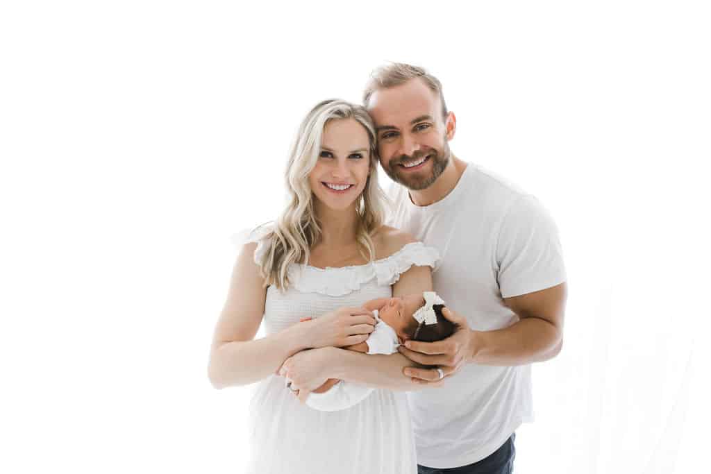 Smiling couple in white clothing holds a newborn baby wrapped in a white blanket against a white background.