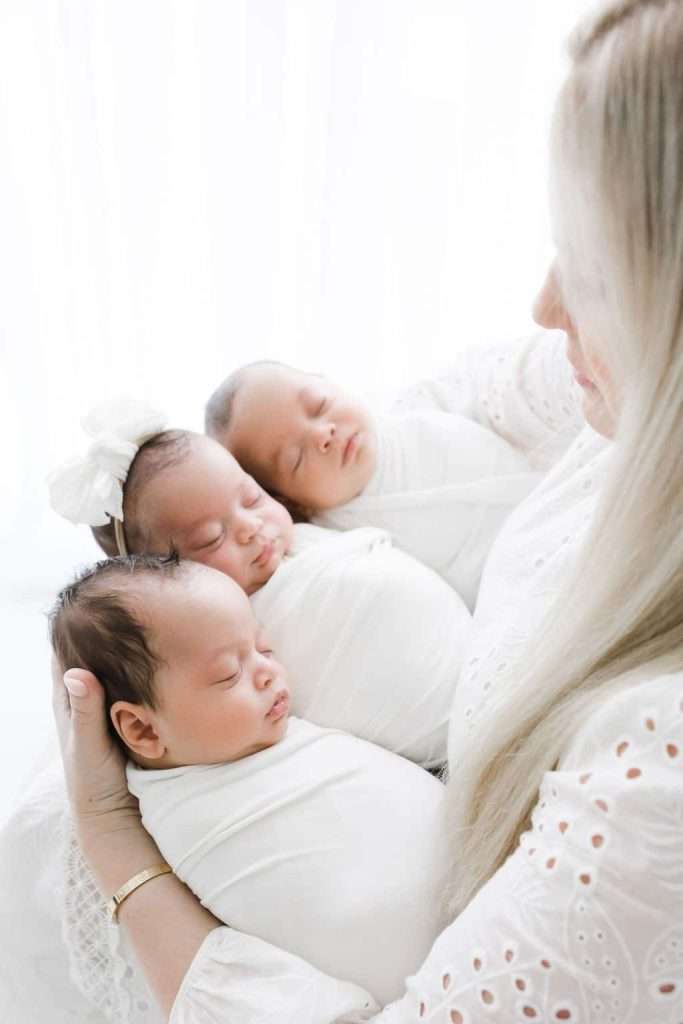 A woman in white holds three swaddled newborn babies, two girls and one boy, as they sleep. The background is bright and softly lit.