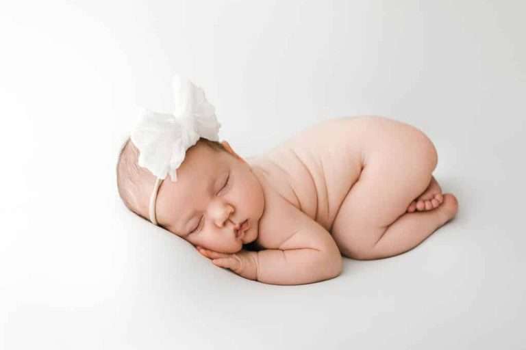 A newborn baby wearing a white headband with a large bow sleeps on their side against a plain white background.