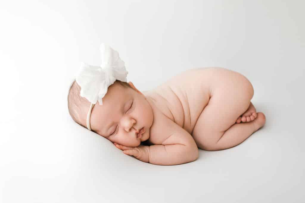 A newborn baby wearing a white headband with a large bow sleeps on their side against a plain white background.
