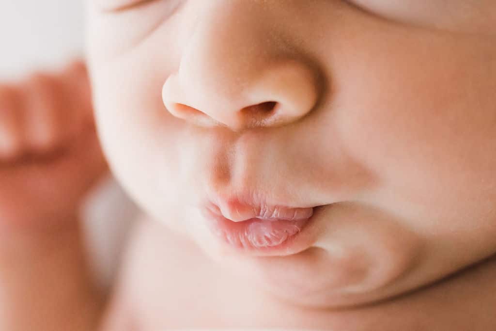 Close-up of a baby’s face showing pursed lips and part of the nose and cheek, with a small fist visible in the background.