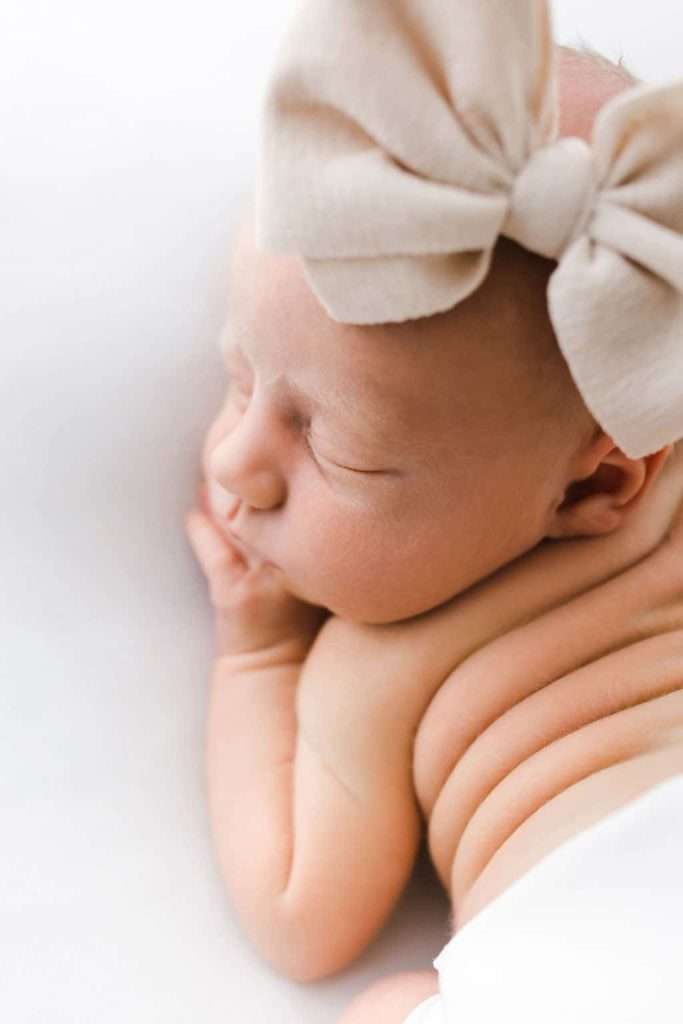 Newborn baby sleeping on their side, wearing a large soft bow headband, with visible baby rolls and eyes closed.