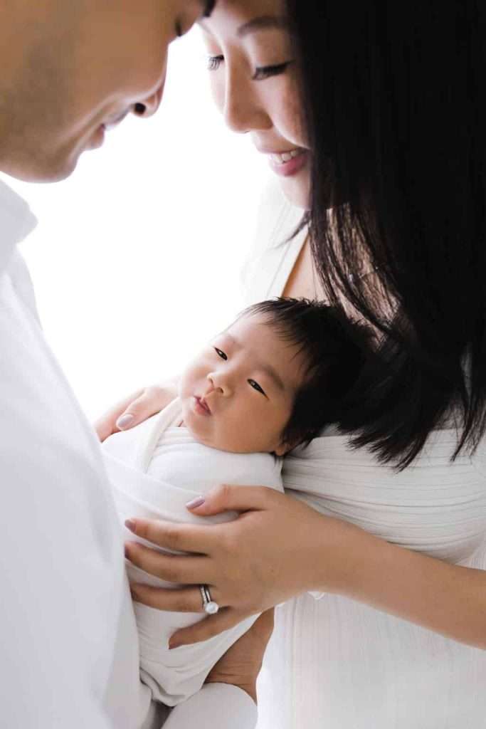A couple holds a newborn baby wrapped in a white blanket. The baby looks forward while the parents smile and gaze at the child.