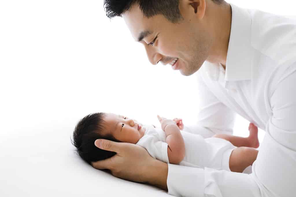 An adult gently holds and looks down at a newborn baby, both wearing white, against a bright, white background.
