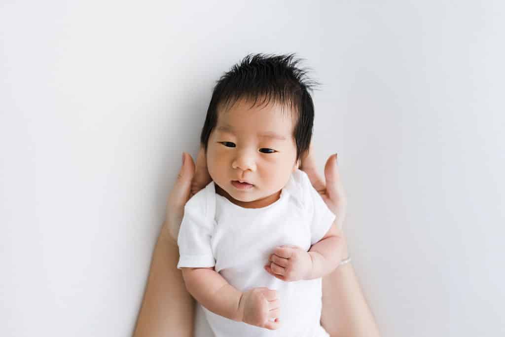 A baby in a white onesie lies on a white surface while an adult’s hands gently support the baby's head and back.