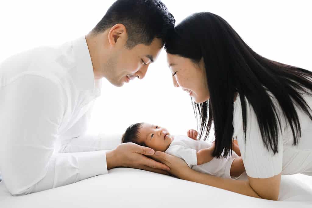 Two adults lean their foreheads together, smiling at a baby lying between them. The baby looks up, supported gently by the adults' hands. All wear white clothing against a bright background.