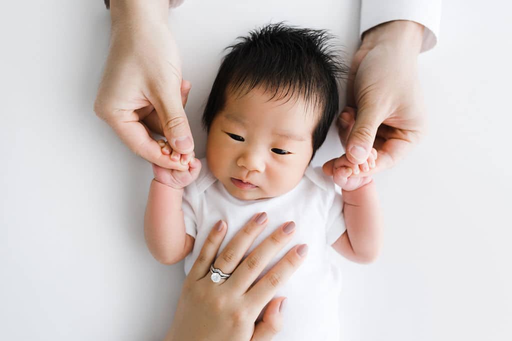 Newborn baby in a white outfit lies on a white surface, with two adult hands gently holding the baby's arms and one hand resting on the baby's chest.